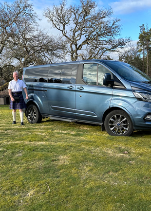 man in a kilt standing beside a blue van on green grass with trees in background suitable for 4 adventures or 4 friends